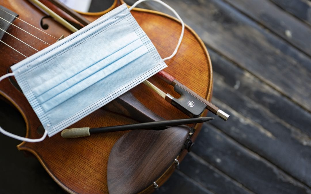 A face mask on an acoustic violin or viola with a pencil and a bow next to it representing performance restrictions during a pandemic